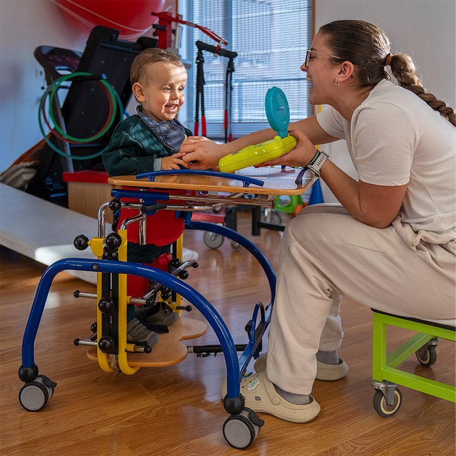 Trabajadora_de_astrapace_con_niño_en_un_bipedestador_sonriendo_mientras_mira_un_juguete